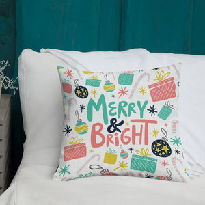 A white pillow with illustrations leading on white bedding with a side table off to the side. The white pillow features illustrated presents, candy canes, stars and ornaments. The middle of the pillow has the words Merry & Bright with the Christmas pattern around it. The colors of the pattern and words are pink, yellow, light blue and black. 
