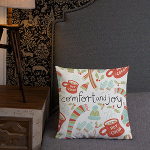 A pillow leaning on a grey headboard with a table and lamp off to the side. The white pillow features Christmas illustrations with scarves, winter hats, mittens, hot cocoa mugs and holy leaves. The words Comfort & Joy are in the center of the pillow in black. The colors of the pattern are in red, light blue and light green. 