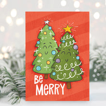 Load image into Gallery viewer, A Christmas card standing up with with pine leaves in the background with a touch of snow. The Christmas card has a red background with two illustrated cute Christmas trees and the words &quot;Be Merry&quot; in white.