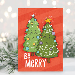 A Christmas card standing up with with pine leaves in the background with a touch of snow. The Christmas card has a red background with two illustrated cute Christmas trees and the words "Be Merry" in white.