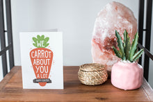 Load image into Gallery viewer, A card on a wood tabletop and on the right side of the card is a woven basket, a pink plant pot with a cactus in it and a pink crystal rock. The card features the words “We carrot about you a lot, Happy Easter,” all featured in an illustrated carrot.