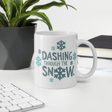 Load image into Gallery viewer, A mug featured on a desk with a plant and a keyboard. The mug is white with the very top silver enamel. The design features the words &quot;Dashing through the snow&quot; in dark and light blue. There are snowflakes around the words. 