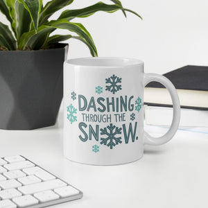 A mug featured on a desk with a plant and a keyboard. The mug is white with the very top silver enamel. The design features the words "Dashing through the snow" in dark and light blue. There are snowflakes around the words. 