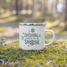 Load image into Gallery viewer, A white enamel mug with a silver enamel rim sitting in the grass. The illustrated design says &quot;Dashing throug the snow&quot; with the &quot;O&quot; of snow as a snowflake. There are snowflakes surrounding the words. The words and snowflakes are in dark and light blue. 