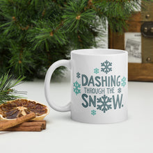 Load image into Gallery viewer, A white mug with a pine tree in the background. The design is in light and dark blue and reads &quot;Dashing through the snow&quot; with snowflakes around the words. 