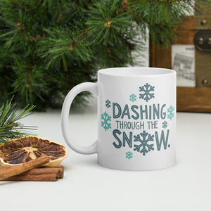 A white mug with a pine tree in the background. The design is in light and dark blue and reads "Dashing through the snow" with snowflakes around the words. 