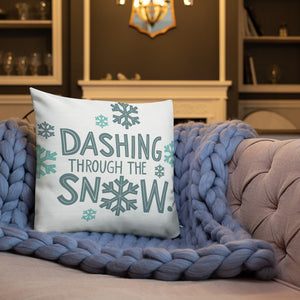 A white pillow on a sofa with a blue knitted blanket. The white pillow features the words "Dashing through the snow" in a repeat pattern with snowflakes. All the letters and illustrations are in lright and dark blue. 