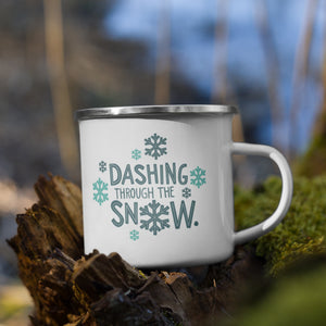 White enamel mug featured on a white table top and white background. The design is in light and dark blue and reads "Dashing through the snow" with snowflakes around the words. 