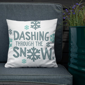 The white pillow is leaning on a sofa with a plant off to the side. The pillow is white and has the words in light and dark blue saying "Dashing through the snow." The words are repeated in a pattern with snowflakes all over the pillow. 