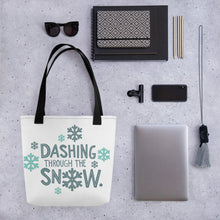 Load image into Gallery viewer, A tote bag lying on a surface with a laptop and office items next to it. The tote bag has the song lyrics &quot;Dashing through the snow&quot; in light and dark blue along with snowflakes surrounding the lettering.  