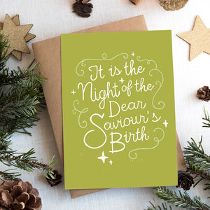 A photo of a Christmas card on top of a brown paper wrapped gift with Christmas decor around it.  The background of the card is a lime green with the word "it is the night of the dear saviour's birth" in script white lettering. 