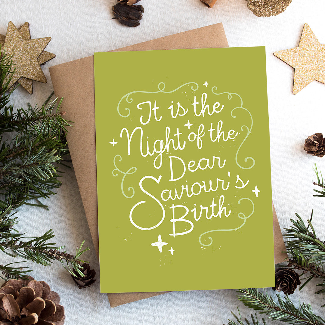 A photo of a Christmas card on top of a brown paper wrapped gift with Christmas decor around it.  The background of the card is a lime green with the word 