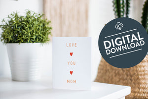 A greeting card is featured on a white tabletop with a white planter in the background with a green plant. There’s a woven basket in the background with a cactus inside. The card features the words ”Love You Mom.” The words "digital download" are featured in a circle on top of the image. 