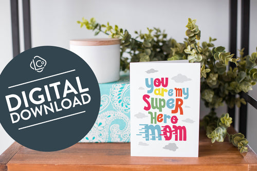 A greeting card is on a table top with a present in blue wrapping paper in the background. On top of the present is a candle and some greenery from a plant too. The card features the words “You are my super hero mom.” The words 