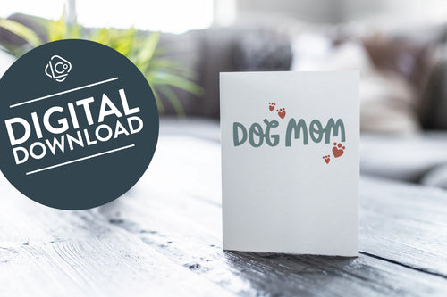 A greeting card featured on a black, wood coffee table. There’s a white planter in the background with a green plant. There’s also a gray sofa in the background with a white pillow. The card features the words “Dog mom.” The words 