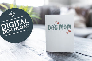 A greeting card featured on a black, wood coffee table. There’s a white planter in the background with a green plant. There’s also a gray sofa in the background with a white pillow. The card features the words “Dog mom.” The words "digital download" are featured in a circle on top of the image. 