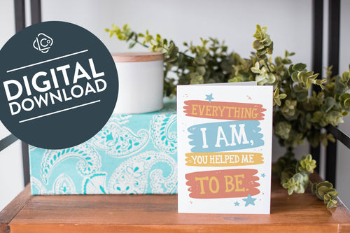A greeting card is on a table top with a present in blue wrapping paper in the background. On top of the present is a candle and some greenery from a plant too. The card features the words “Everything I Am, You Helped Me To Be.” The words 