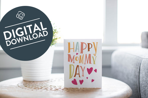 A greeting card is featured on a desktop with a green plant to the side. The card features a couple illustrated hearts with the words “Happy Mommy Day.” The words 