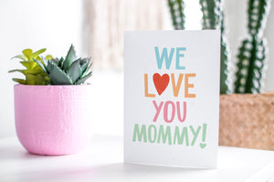 A greeting card featured standing up on a white tabletop with a pink plant pot in the background and some succulents in the pot. There’s a woven basket in the background with a cactus inside. The card features the words “We Love You Mommy!” 