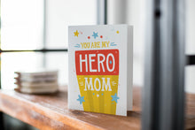 Load image into Gallery viewer, A card on a wood tabletop with an object in the background that is out of focus. The card features the words “You are my hero mom.”