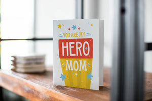 A card on a wood tabletop with an object in the background that is out of focus. The card features the words “You are my hero mom.”