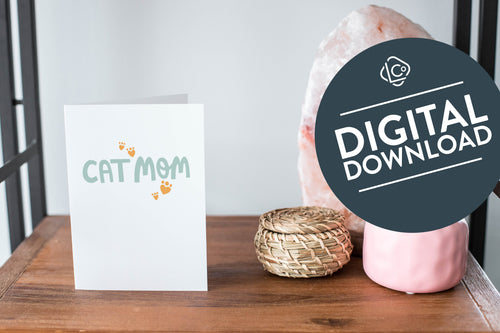 A card on a wood tabletop and on the right side of the card is a woven basket, a pink plant pot with a cactus in it and a pink crystal rock. The card features the words “Cat Mom.” The words 