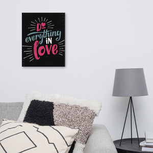 A black art canvas hangs on a white living room wall above a grey sofa. The canvas reads "Do everything in love" in bright pink and blue hand-lettering style, with white dashes around the words. The sofa is covered with black and white cushions, with a grey table lamp to the right.