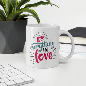 A white mug sits on a white desk, next to a computer keyboard, potted plant and notebooks. The mug reads "Do everything in love' in bright pink and blue lettering, with black dashes coming out from the words.