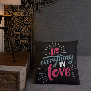 A square black pillow sits on a grey bedspread in front of a wallpapered wall. The pillow reads "Do everything in love" in bright pink and blue hand-lettering style, with white dashes around the words.
