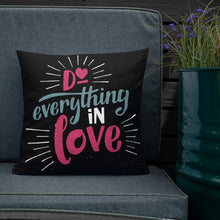 Load image into Gallery viewer, A square black pillow sits on a grey sofa beside a planter of purple flowers. The pillow reads &quot;Do everything in love&quot; in bright pink and blue hand-lettering style, with white dashes around the words.