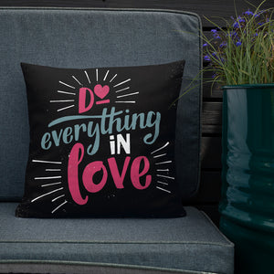 A square black pillow sits on a grey sofa beside a planter of purple flowers. The pillow reads "Do everything in love" in bright pink and blue hand-lettering style, with white dashes around the words.