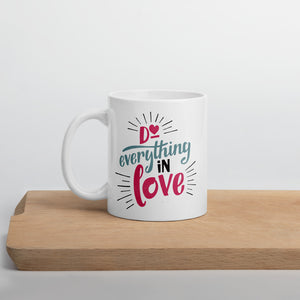 A white mug sits on a wooden chopping board against a white wall. The mug reads "Do everything in love' in bright pink and blue lettering, with black dashes coming out from the words.