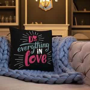 A square black pillow sits on a pink sofa covered with a chunky lilac blanket. The pillow reads "Do everything in love" in bright pink and blue hand-lettering style, with white dashes around the words.