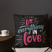 Load image into Gallery viewer, A square black pillow sits on a chair in front of a grey wall. The pillow reads &quot;Do everything in love&quot; in bright pink and blue hand-lettering style, with white dashes around the words.