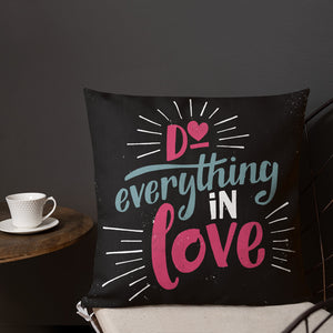 A square black pillow sits on a chair in front of a grey wall. The pillow reads "Do everything in love" in bright pink and blue hand-lettering style, with white dashes around the words.