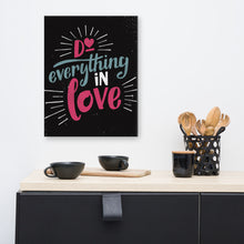 Load image into Gallery viewer, A black art canvas hangs on a white kitchen wall, above a black sideboard. The canvas reads &quot;Do everything in love&quot; in bright pink and blue hand-lettering style, with white dashes around the words. On the sideboard sit black cups and wooden utensils.