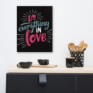 A black art canvas hangs on a white kitchen wall, above a black sideboard. The canvas reads "Do everything in love" in bright pink and blue hand-lettering style, with white dashes around the words. On the sideboard sit black cups and wooden utensils.
