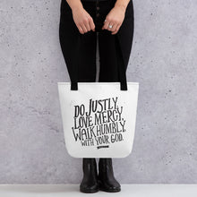 Load image into Gallery viewer, A tote bag with black handles and white fabric bag. The bag features black lettering in the words Do justly, love mercy, walk humbly, with your God, Micah 6:8.