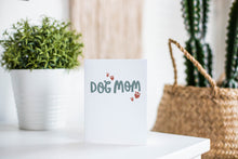 Load image into Gallery viewer, A greeting card is featured on a white tabletop with a white planter in the background with a green plant. There’s a woven basket in the background with a cactus inside. The card features the words ”Dog Mom.”