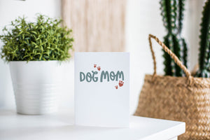 A greeting card is featured on a white tabletop with a white planter in the background with a green plant. There’s a woven basket in the background with a cactus inside. The card features the words ”Dog Mom.”