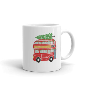 A white ceramic mug featured on a white background. The mug features the traditional London bus, a red double decker bus. The illustration also has a Christmas tree laying on the top and green Christmas garland in the windows of the bus. 