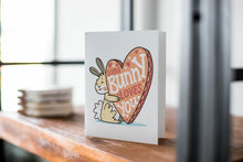 Load image into Gallery viewer, A card on a wood tabletop with an object in the background that is out of focus. The card features an illustrated Easter bunny holding a heart with the words “some bunny loves you.” 
