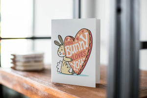 A card on a wood tabletop with an object in the background that is out of focus. The card features an illustrated Easter bunny holding a heart with the words “some bunny loves you.” 