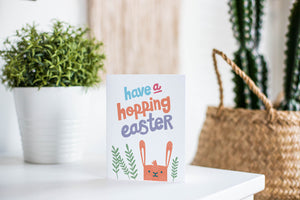 A greeting card is featured on a white tabletop with a white planter in the background with a green plant. There’s a woven basket in the background with a cactus inside. The card features an illustrated Easter bunny with some leaves around the bunny. The words “have a hopping Easter” are above the bunny. 