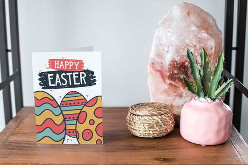 A card on a wood tabletop and on the right side of the card is a woven basket, a pink plant pot with a cactus in it and a pink crystal rock. The card features the words “Happy Easter” with illustrated Easter eggs.