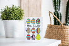 Load image into Gallery viewer, A greeting card is featured on a white tabletop with a white planter in the background with a green plant. There’s a woven basket in the background with a cactus inside. The card features illustrated Easter eggs in bright fun colors with the words “Happy Easter” in the middle of the eggs. 