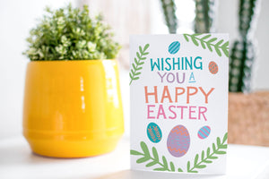 A greeting card is on a table top with a yellow plant pot and a green plant inside. The card features the words “Wishing you a happy Easter” with illustrated Easter eggs and palm leaves. 