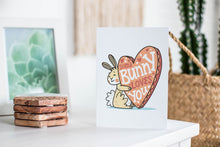 Load image into Gallery viewer, A greeting card featured standing up on a white tabletop with a framed photo of a succulent in the background and a stack of wooden coasters. There’s a woven basket in the background with a cactus inside. The card features an illustrated Easter bunny holding a heart with the words “some bunny loves you.”