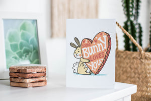 A greeting card featured standing up on a white tabletop with a framed photo of a succulent in the background and a stack of wooden coasters. There’s a woven basket in the background with a cactus inside. The card features an illustrated Easter bunny holding a heart with the words “some bunny loves you.”