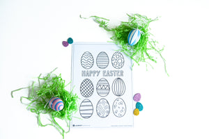 A coloring sheet on a white tabletop. There’s fake Easter grass around the color page. The coloring page features illustrated Easter eggs with fun patterns to color in with the words “Happy Easter” in the middle of the eggs. 